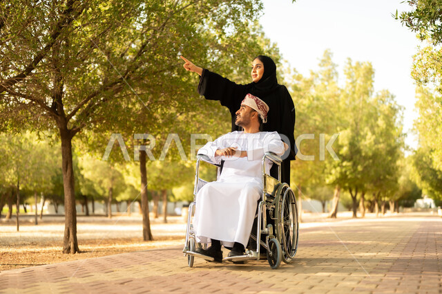 الاستمتاع وقضاء وقت ممتع بالهواء الطلق, العطف والحب بين أفراد الأسرة, امرأة عربية خليجية عمانية محجبة ترتدي العباءة تتنزه في الحديقة برفقة زوجها المقعد, شعور ذوي الهمم بالراحة والطمأنينة في بيئتهم, مساعدة مرضى الإعاقة الحركية على التنقل بسهولة