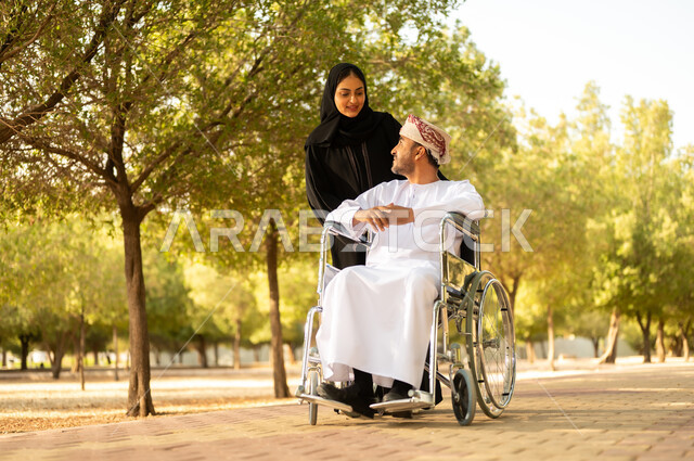 العطف والحب بين أفراد الأسرة, امرأة عربية خليجية عمانية محجبة ترتدي العباءة تتنزه في الحديقة برفقة زوجها المقعد, شعور ذوي الهمم بالراحة والطمأنينة في بيئتهم, مساعدة مرضى الإعاقة الحركية على التنقل بسهولة، الاستمتاع وقضاء وقت ممتع بالهواء الطلق