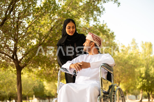 الاستمتاع وقضاء وقت ممتع بالهواء الطلق, العطف والحب بين أفراد الأسرة, امرأة عربية خليجية عمانية محجبة ترتدي العباءة تتنزه في الحديقة برفقة زوجها المقعد, شعور ذوي الهمم بالراحة والطمأنينة في بيئتهم, مساعدة مرضى الإعاقة الحركية على التنقل بسهولة