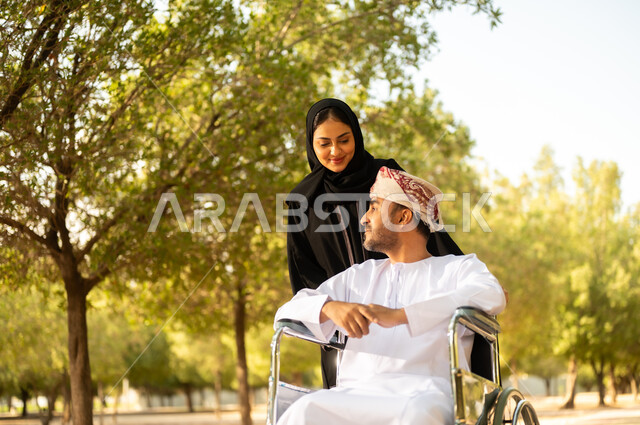 مساعدة مرضى الإعاقة الحركية على التنقل بسهولة، الاستمتاع وقضاء وقت ممتع بالهواء الطلق، العطف والحب بين أفراد الأسرة, امرأة عربية خليجية عمانية محجبة ترتدي العباءة تتنزه في الحديقة برفقة زوجها المقعد, شعور ذوي الهمم بالراحة والطمأنينة في بيئتهم
