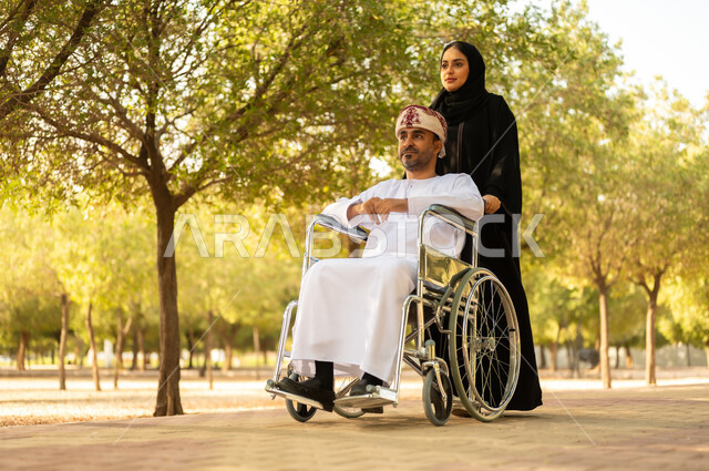 العطف والحب بين أفراد الأسرة, امرأة عربية خليجية عمانية محجبة ترتدي العباءة تتنزه في الحديقة برفقة زوجها المقعد, شعور ذوي الهمم بالراحة والطمأنينة في بيئتهم, مساعدة مرضى الإعاقة الحركية على التنقل بسهولة، الاستمتاع وقضاء وقت ممتع بالهواء الطلق