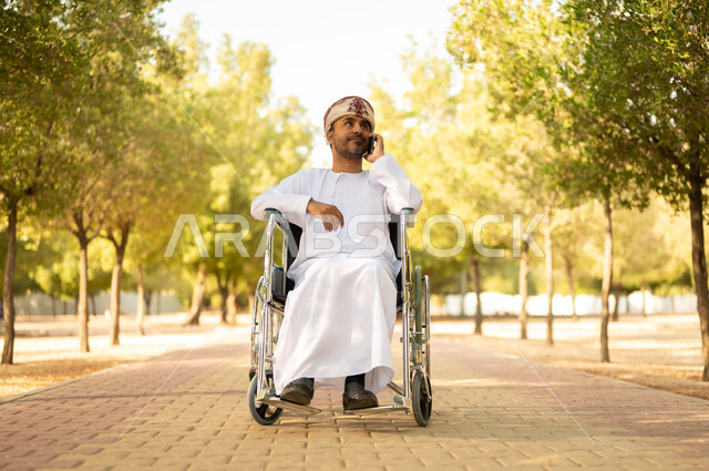 نمط حياة ذوي الهمم في سلطنة عمان, رجل مقعد عربي خليجي عامني يرتدي الدشداشة والعمامة يجلس على كرسي متحرك يجري مكالمة هاتفيه عبر الهاتف المحمول، التنزه في إحدى الحدائق العامة، قضاء وقت ممتع في المنتزه في النهار، استخدام التقنية الحديثة في الهواء الطلق