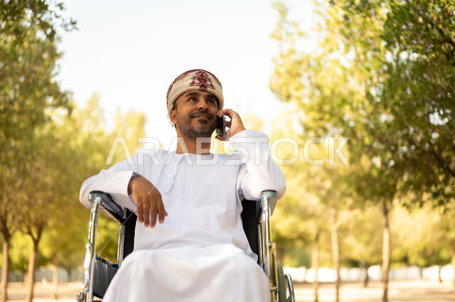 نمط حياة ذوي الهمم في سلطنة عمان, رجل مقعد عربي خليجي عامني يرتدي الدشداشة والعمامة يجلس على كرسي متحرك يجري مكالمة هاتفيه عبر الهاتف المحمول، التنزه في إحدى الحدائق العامة، قضاء وقت ممتع في المنتزه في النهار، استخدام التقنية الحديثة في الهواء الطلق