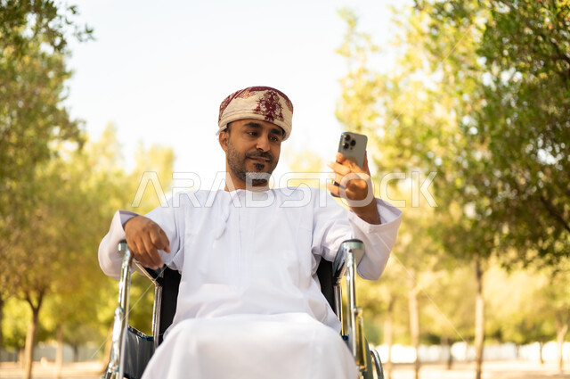 قضاء وقت ممتع في المنتزه في النهار، استخدام التقنية الحديثة في الهواء الطلق, نمط حياة ذوي الهمم في سلطنة عمان, رجل مقعد عربي خليجي عامني يرتدي الدشداشة والعمامة يجلس على كرسي متحرك يمسك بيده الهاتف المحمول، التنزه في إحدى الحدائق العامة