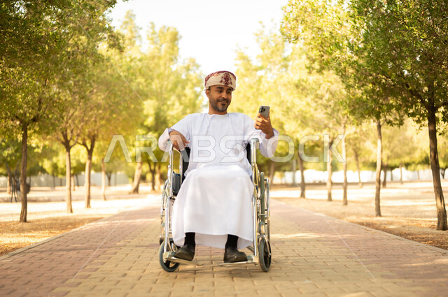 قضاء وقت ممتع في المنتزه في النهار، استخدام التقنية الحديثة في الهواء الطلق, نمط حياة ذوي الهمم في سلطنة عمان, رجل مقعد عربي خليجي عامني يرتدي الدشداشة والعمامة يجلس على كرسي متحرك يمسك بيده الهاتف المحمول، التنزه في إحدى الحدائق العامة