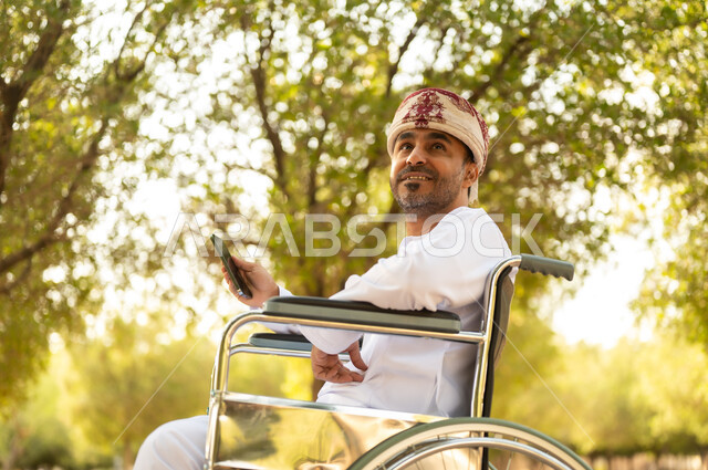 استخدام التقنية الحديثة في الهواء الطلق, نمط حياة ذوي الهمم في سلطنة عمان, رجل مقعد عربي خليجي عامني يرتدي الدشداشة والعمامة يجلس على كرسي متحرك يمسك بيده الهاتف المحمول، التنزه في إحدى الحدائق العامة, قضاء وقت ممتع في المنتزه في النهار