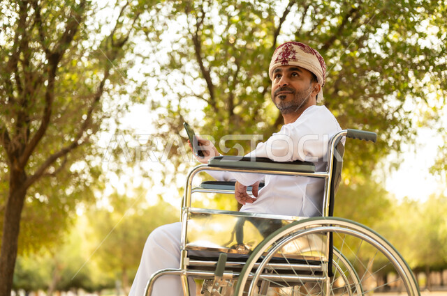 استخدام التقنية الحديثة في الهواء الطلق, نمط حياة ذوي الهمم في سلطنة عمان, رجل مقعد عربي خليجي عامني يرتدي الدشداشة والعمامة يجلس على كرسي متحرك يمسك بيده الهاتف المحمول، التنزه في إحدى الحدائق العامة, قضاء وقت ممتع في المنتزه في النهار
