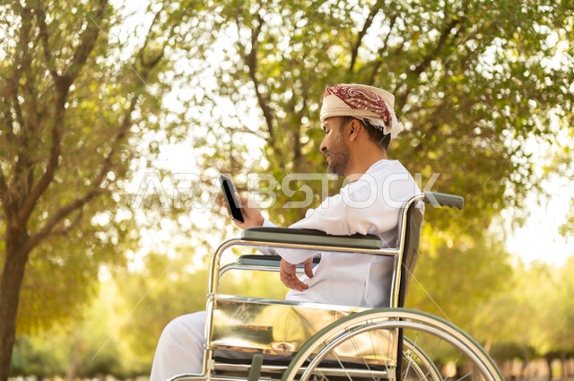 قضاء وقت ممتع في المنتزه في النهار، استخدام التقنية الحديثة في الهواء الطلق, نمط حياة ذوي الهمم في سلطنة عمان, رجل مقعد عربي خليجي عامني يرتدي الدشداشة والعمامة يجلس على كرسي متحرك يمسك بيده الهاتف المحمول، التنزه في إحدى الحدائق العامة