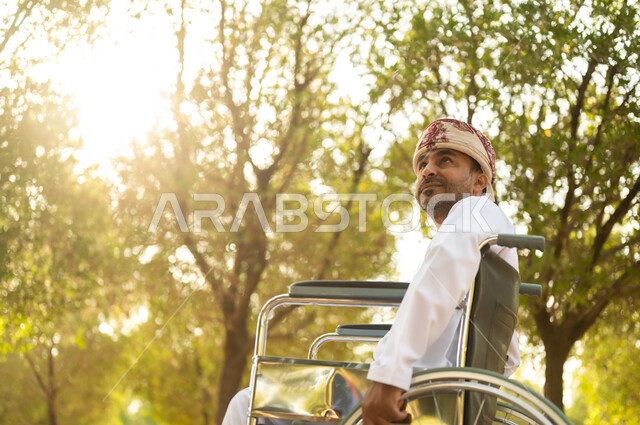 الاستمتاع بالهواء الطلق, نمط حياة ذوي الهمم في سلطنة عمان, رجل مقعد عربي خليجي عامني يرتدي الدشداشة والعمامة يجلس على كرسي متحرك ويتنزه في إحدى الحدائق العامة, قضاء وقت ممتع في المنتزه في النهار
