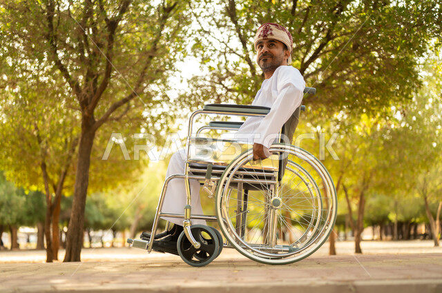 الاستمتاع بالهواء الطلق, نمط حياة ذوي الهمم في سلطنة عمان, رجل مقعد عربي خليجي عامني يرتدي الدشداشة والعمامة يجلس على كرسي متحرك ويتنزه في إحدى الحدائق العامة, قضاء وقت ممتع في المنتزه في النهار