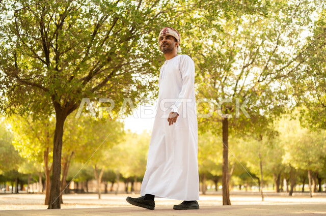 جولة في الهواء الطلق, قضاء أوقات ممتعة في الحدائق الخضراء، رجل عربي خليجي عماني يرتدي الدشداشة والعمامة يتنزه في الحديقة العامة في النهار، إيماءات وتعابير السرور والسعادة, خلفية الطبيعة