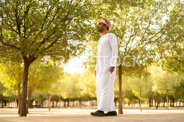 جولة في الهواء الطلق, قضاء أوقات ممتعة في الحدائق الخضراء، رجل عربي خليجي عماني يرتدي الدشداشة والعمامة يتنزه في الحديقة العامة في النهار، إيماءات وتعابير السرور والسعادة, خلفية الطبيعة