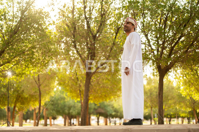 جولة في الهواء الطلق, قضاء أوقات ممتعة في الحدائق الخضراء، رجل عربي خليجي عماني يرتدي الدشداشة والعمامة يتنزه في الحديقة العامة في النهار، إيماءات وتعابير السرور والسعادة, خلفية الطبيعة