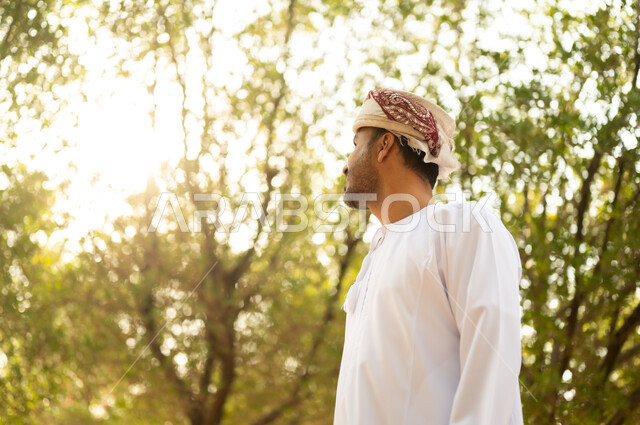 جولة في الهواء الطلق, قضاء أوقات ممتعة في الحدائق الخضراء، رجل عربي خليجي عماني يرتدي الدشداشة والعمامة يتنزه في الحديقة العامة في النهار، إيماءات وتعابير السرور والسعادة, خلفية الطبيعة