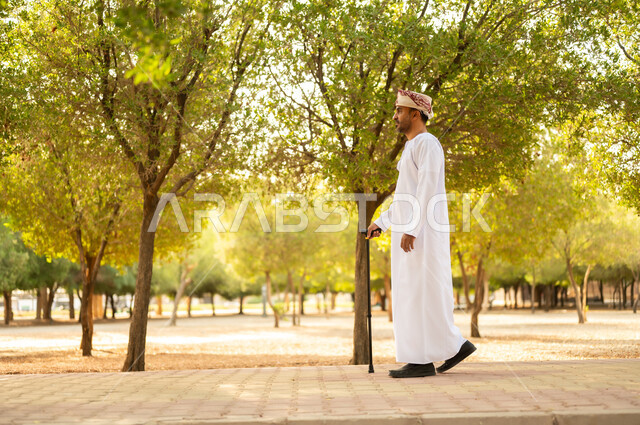 جودة حياة كبار السن في سلطنة عمان، التجول في الطبيعة واستنشاق الهواء الطلق، الاستمتاع بالأجواء الصيفيه في إحدى المنتزهات، رجل عربي خليجي عماني يرتدي الدشداشة والعمامة يرتكز على العكاز لسهولة التنقل، معدات تساعد على المشي