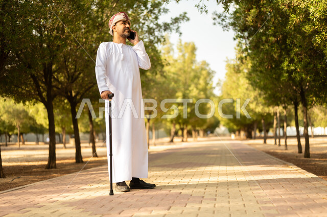 اجراء مكالمة هاتفية عبر الهاتف المحمول، استخدام جهاز تقني حديث، جودة حياة كبار السن في سلطنة عمان، التجول في الطبيعة واستنشاق الهواء الطلق، الاستمتاع بالأجواء الصيفيه في المنتزه، رجل عربي خليجي عماني يرتدي الدشداشة والعمامة يرتكز على العكاز لسهولة التنقل