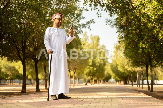 استخدام جهاز تقني حديث، جودة حياة كبار السن في سلطنة عمان، التجول في الطبيعة واستنشاق الهواء الطلق، الاستمتاع بالأجواء الصيفيه في المنتزه، رجل عربي خليجي عماني يرتدي الدشداشة والعمامة يمسك بيده الهاتف المحمول يرتكز على العكاز لسهولة التنقل