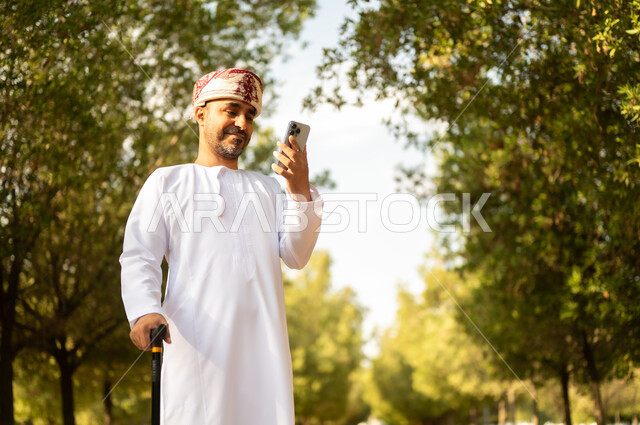 استخدام جهاز تقني حديث، جودة حياة كبار السن في سلطنة عمان، التجول في الطبيعة واستنشاق الهواء الطلق، الاستمتاع بالأجواء الصيفيه في المنتزه، رجل عربي خليجي عماني يرتدي الدشداشة والعمامة يمسك بيده الهاتف المحمول يرتكز على العكاز لسهولة التنقل
