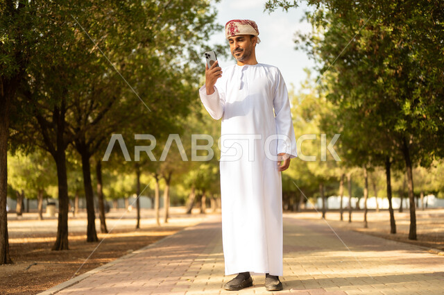 استخدام جهاز تقني حديث، قضاء أوقات ممتعة في الحدائق الخضراء, جولة في الهواء الطلق, رجل عربي خليجي عماني يرتدي الدشداشة والعمامة يمسك بيده الهاتف المحمول يتنزه في الحديقة العامة في النهار، إيماءات وتعابير السرور والسعادة, خلفية الطبيعة