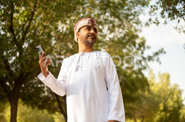 استخدام جهاز تقني حديث، قضاء أوقات ممتعة في الحدائق الخضراء, جولة في الهواء الطلق, رجل عربي خليجي عماني يرتدي الدشداشة والعمامة يمسك بيده الهاتف المحمول يتنزه في الحديقة العامة في النهار، إيماءات وتعابير السرور والسعادة, خلفية الطبيعة