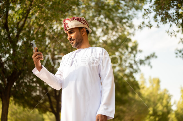 استخدام جهاز تقني حديث، قضاء أوقات ممتعة في الحدائق الخضراء, جولة في الهواء الطلق, رجل عربي خليجي عماني يرتدي الدشداشة والعمامة يمسك بيده الهاتف المحمول يتنزه في الحديقة العامة في النهار، إيماءات وتعابير السرور والسعادة, خلفية الطبيعة