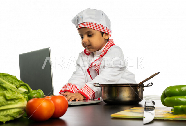Portrait of a Saudi Arabian Gulf boy, dressed in a special chef uniform, standing in front of a table with a group of fresh vegetables, using a laptop, sharing preparing recipes on social media, preparing salads and healthy dishes, a future profession