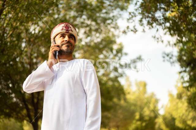 اجراء مكالمة هاتفية، قضاء أوقات ممتعة في الحدائق الخضراء, جولة في الهواء الطلق, رجل عربي خليجي عماني يرتدي الدشداشة والعمامة يمسك بيده الهاتف المحمول يتنزه في الحديقة العامة في النهار، إيماءات وتعابير السرور والسعادة, خلفية الطبيعة
