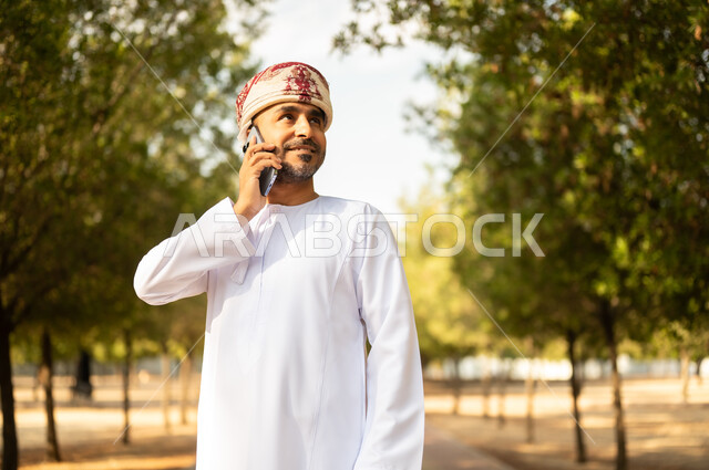 Making a phone call, having fun in green parks, outdoor walk, an Arab Gulf Omani man wearing a dishdasha and turban holding a mobile phone, walking in a public park during the day, gestures and expressions of pleasure and happiness, nature background