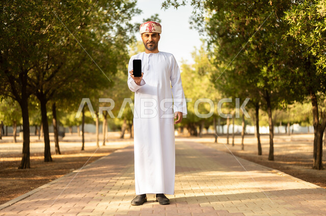 عرض شاشة سوداء فار غة، قضاء أوقات ممتعة في الحدائق الخضراء, جولة في الهواء الطلق, رجل عربي خليجي عماني يرتدي الدشداشة والعمامة يمسك بيده الهاتف المحمول يتنزه في الحديقة العامة في النهار، إيماءات وتعابير السرور والسعادة, خلفية الطبيعة