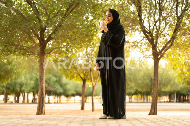 تناول المشروبات الساخنة، شابة عربية خليجية سعودية تتجول في إحدى الحدائق العامة، قضاء أوقات ممتعة، ايماءات وتعابير السعادة و السرور، إمرأة عمانية ترتدي العباءة والحجاب تقوم بنزهه في الهواء الطلق في احدى الحدائق والمنتزهات