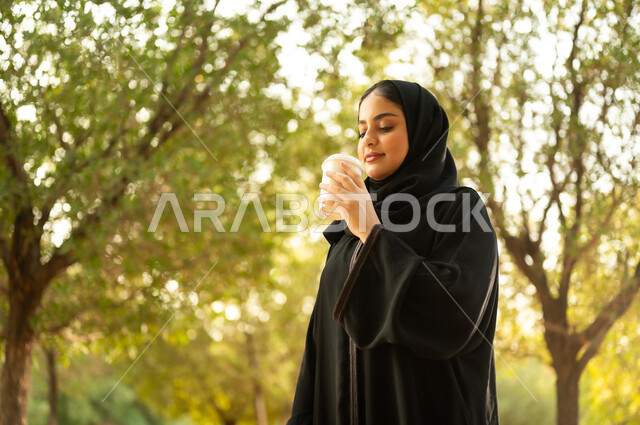 تناول المشروبات الساخنة، شابة عربية خليجية سعودية تتجول في إحدى الحدائق العامة، قضاء أوقات ممتعة، ايماءات وتعابير السعادة و السرور، إمرأة عمانية ترتدي العباءة والحجاب تقوم بنزهه في الهواء الطلق في احدى الحدائق والمنتزهات