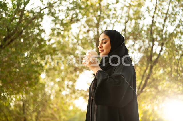 تناول المشروبات الساخنة، شابة عربية خليجية سعودية تتجول في إحدى الحدائق العامة، قضاء أوقات ممتعة، ايماءات وتعابير السعادة و السرور، إمرأة عمانية ترتدي العباءة والحجاب تقوم بنزهه في الهواء الطلق في احدى الحدائق والمنتزهات