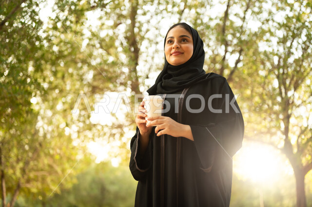 تناول المشروبات الساخنة، شابة عربية خليجية سعودية تتجول في إحدى الحدائق العامة، قضاء أوقات ممتعة، ايماءات وتعابير السعادة و السرور، إمرأة عمانية ترتدي العباءة والحجاب تقوم بنزهه في الهواء الطلق في احدى الحدائق والمنتزهات
