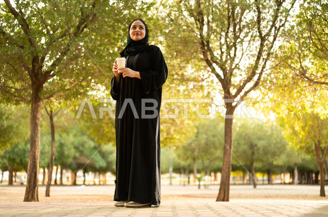 تناول المشروبات الساخنة، شابة عربية خليجية سعودية تتجول في إحدى الحدائق العامة، قضاء أوقات ممتعة، ايماءات وتعابير السعادة و السرور، إمرأة عمانية ترتدي العباءة والحجاب تقوم بنزهه في الهواء الطلق في احدى الحدائق والمنتزهات
