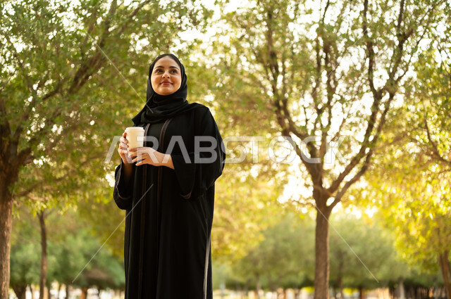 تناول المشروبات الساخنة، شابة عربية خليجية سعودية تتجول في إحدى الحدائق العامة، قضاء أوقات ممتعة، ايماءات وتعابير السعادة و السرور، إمرأة عمانية ترتدي العباءة والحجاب تقوم بنزهه في الهواء الطلق في احدى الحدائق والمنتزهات