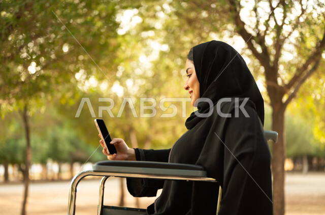 استخدام جهاز تقني تكنولوجي حديث، إمرأة مقعدة عربية خليجية عمانية ترتدي الحجاب والعباءة تمسك بيدها الهاتف المحمول تجلس على كرسي متحرك في احدى المنتزهات، توفير اجهزة ومعدات تساعد بالحركة والتنقل، سيدة سعودية من ذوي الاحتياجات الخاصه تقضي وقت ممتع في الحديقة