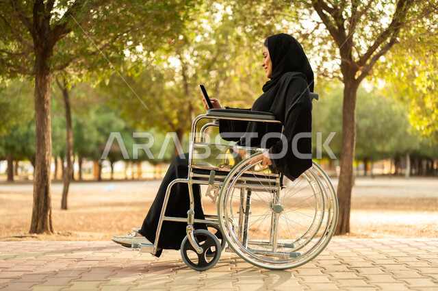استخدام جهاز تقني تكنولوجي حديث، إمرأة مقعدة عربية خليجية عمانية ترتدي الحجاب والعباءة تمسك بيدها الهاتف المحمول تجلس على كرسي متحرك في احدى المنتزهات، توفير اجهزة ومعدات تساعد بالحركة والتنقل، سيدة سعودية من ذوي الاحتياجات الخاصه تقضي وقت ممتع في الحديقة