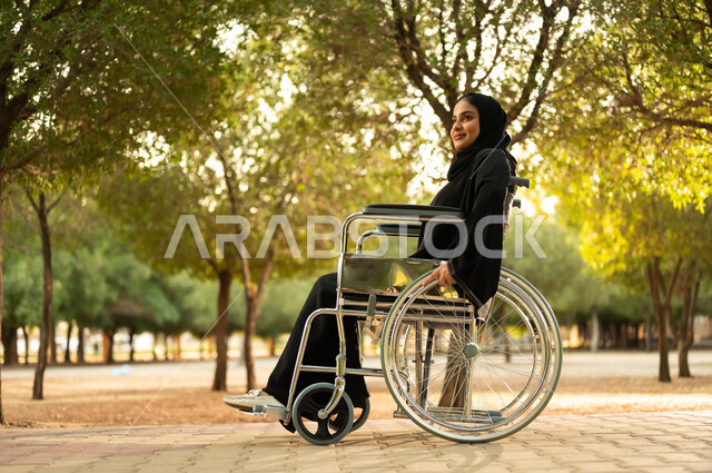 توفير اجهزة ومعدات تساعد بالحركة والتنقل، سيدة سعودية من ذوي الاحتياجات الخاصه تقضي وقت ممتع في الحديقة، شعور المعاقين بالإنتماء والتقدير، إمرأة مقعدة عربية خليجية عمانية ترتدي الحجاب والعباءة تجلس على كرسي متحرك في احدى المنتزهات