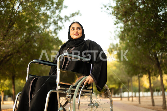 Providing devices and equipment to aid movement and mobility, a Saudi woman with special needs spending a pleasant time in the park, the sense of belonging and appreciation for the disabled, a disabled Arab Gulf Omani woman wearing a hijab and abaya sitting in a wheelchair in a park