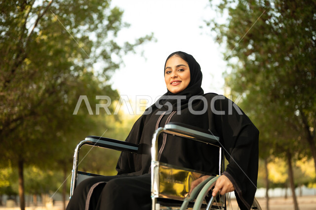 توفير اجهزة ومعدات تساعد بالحركة والتنقل، سيدة سعودية من ذوي الاحتياجات الخاصه تقضي وقت ممتع في الحديقة، شعور المعاقين بالإنتماء والتقدير، إمرأة مقعدة عربية خليجية عمانية ترتدي الحجاب والعباءة تجلس على كرسي متحرك في احدى المنتزهات