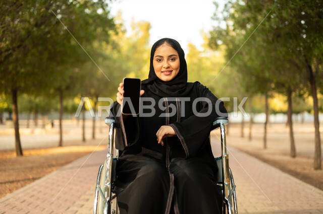 Showing a blank black screen, a disabled Arab Gulf Omani woman wearing a hijab and abaya holding a mobile phone sitting in a wheelchair in a park, providing devices and equipment to help with movement and mobility, a Saudi woman with special needs spending a pleasant time in the park
