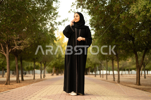 اجراء مكالمة هاتفية، استخدام جهاز تقني حديث، شابة عربية خليجية سعودية تستخدم الهاتف المحمول تتجول في إحدى الحدائق العامة، قضاء أوقات ممتعة، ايماءات وتعابير السعادة و السرور، إمرأة عمانية ترتدي العباءة والحجاب تقوم بنزهه في الهواء الطلق في المنتزه