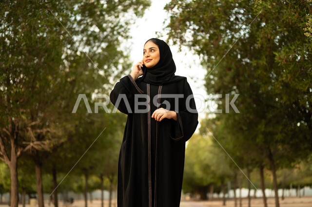 اجراء مكالمة هاتفية، استخدام جهاز تقني حديث، شابة عربية خليجية سعودية تستخدم الهاتف المحمول تتجول في إحدى الحدائق العامة، قضاء أوقات ممتعة، ايماءات وتعابير السعادة و السرور، إمرأة عمانية ترتدي العباءة والحجاب تقوم بنزهه في الهواء الطلق في المنتزه