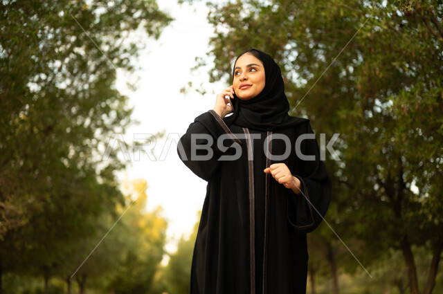اجراء مكالمة هاتفية، استخدام جهاز تقني حديث، شابة عربية خليجية سعودية تستخدم الهاتف المحمول تتجول في إحدى الحدائق العامة، قضاء أوقات ممتعة، ايماءات وتعابير السعادة و السرور، إمرأة عمانية ترتدي العباءة والحجاب تقوم بنزهه في الهواء الطلق في المنتزه