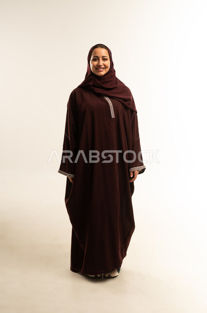 الاناقة والاهتمام بالمظهر الخارجي، ايماءات الثقة بالنفس ، مفهوم الأنوثة والملامح الناعمة, بورتريه لامراة عربية خليجية عمانية ترتدي العباءة والحجاب تقف باستقامة ، سيدة سعودية محجبة تنظر الى الكاميرا بتعابير السعادة, الجسم بالطول الكامل، خلفية بيضاء