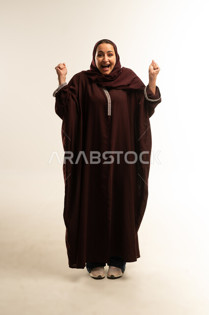 الانتصار والفوز والنجاح، مفهوم الانجاز وتحقيق الاهداف، بورتريه لامراة عربيه خليجية عمانية ترتدي العباءة السوداء تنظرالي الكاميرا و ترفع قبضة يديها للاعلى بإيماءات الفرح والسرور, سيده سعوديه تشعربالسعادة والقوة، صورة الجسم بالطول الكامل, خلفية بيضاء