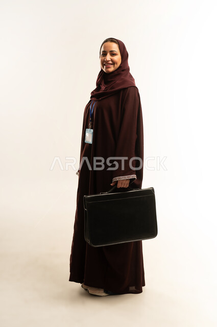 الاهتمام بالمظهر الخارجي،مفهوم إداة الأعمال والمشاريع، بورتريه لامراة عربية خليجية عمانية محجبة ترتدي عباءة بنيه تحمل بيدها حقيبه سوداء، سيده سعودية تنظر الى الكاميرا بايماءات الثقة بالنفس ، ارتداء هوية شخصية، الجسم بالطول الكامل، خلفية بيضاء