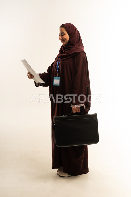 متابعة الخطط والمشاريع،ارتداء هوية شخصية، الأناقة والاهتمام بالمظهر الخارجي، بورتريه لامراة عربية خليجية عمانية محجبة ترتدي عباءة بنيه تحمل بيدها حقيبه سوداء، سيده سعودية تنظر الى مجموعه من الاوراق بايماءات السرور، الجسم بالطول الكامل، خلفية بيضاء