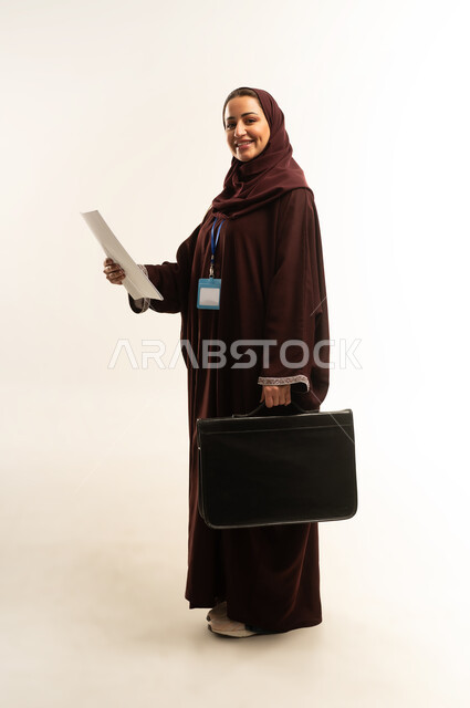 متابعة الخطط والمشاريع، ارتداء هوية شخصية، الأناقة والاهتمام بالمظهر الخارجي، بورتريه لامراة عربية خليجية عمانية محجبة ترتدي عباءة بنيه تحمل بيدها حقيبه سوداء ومجموعه من الاوراق، سيده سعودية تنظر الى شيء ما بايماءات السرور، الجسم بالطول الكامل، خلفية بيضاء