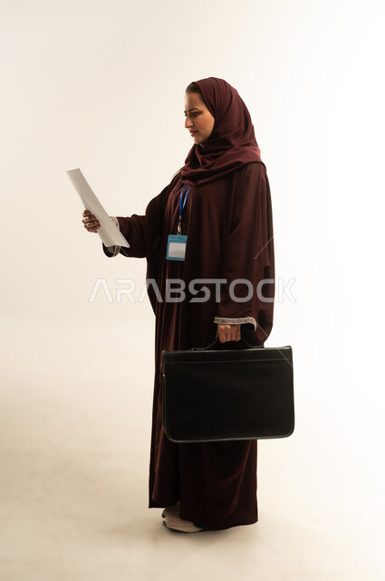 ارتداء هوية شخصية، الأناقة والاهتمام بالمظهر الخارجي، مفهوم إدارة الأعمال والمشاريع، بورتريه لامراة عربية خليجية عمانية محجبة ترتدي عباءة بنيه تحمل بيدها حقيبه سوداء، سيده سعودية تنظر الى مجموعه من الاوراق بايماءات السرور، الجسم بالطول الكامل، خلفية بيضاء